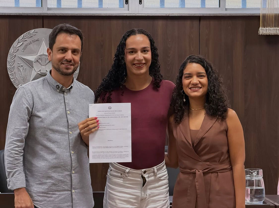 #ParaTodosVerem: Três pessoas centralizadas na foto. À esquerda, Juliano Duarte veste uma camisa social cinza. Ao seu lado, no centro, Yza Fidelis veste uma camiseta bordô e segura um documento voltado para a câmera. A sua direita, a juíza Ana Carolina Ferreira Marques dos Prazeres veste uma blusa marrom. Todos estão sorrindo para a câmera. Ao fundo, uma parede marrom com o selo do estado de Minas Gerais.