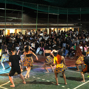 Grupo de jovens realiza apresentação de dança em quadra esportiva à noite, diante de uma grande plateia atenta. Os dançarinos, em sua maioria mulheres, usam saias coloridas com estampas africanas e estão descalços. A iluminação destaca o centro da quadra, onde ocorre a performance. Ao fundo, dezenas de pessoas — de pé e sentadas — assistem à apresentação com expressões de curiosidade e entusiasmo. A cena transmite cultura, movimento e celebração comunitária.