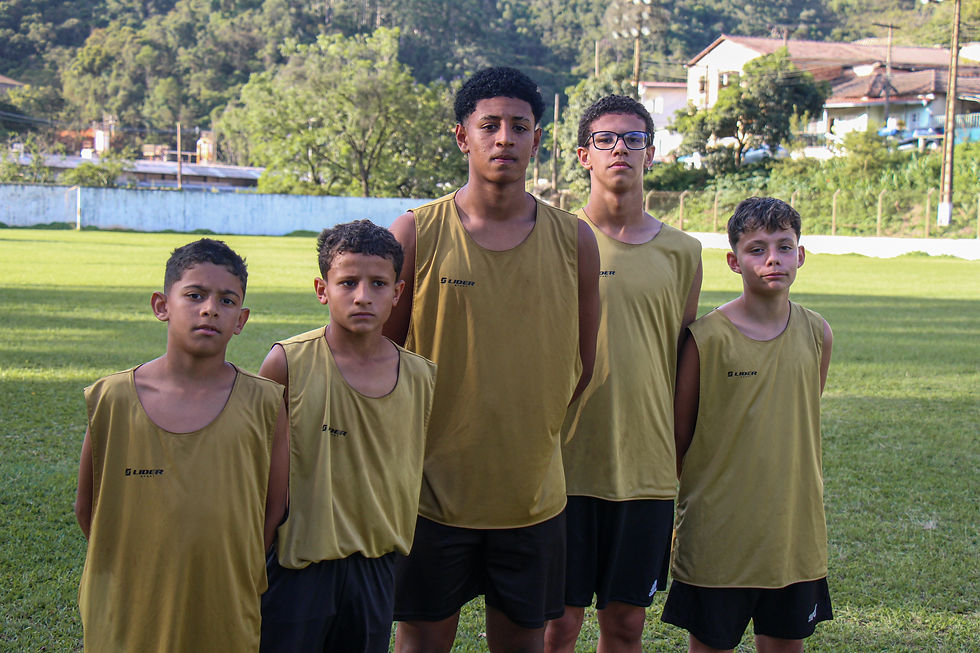 #ParaTodosVerem: Cinco meninos alinhados em pé no gramado de um campo de futebol, usando coletes beges e shorts pretos. Eles olham diretamente para a foto com expressões sérias. Ao fundo, há árvores e casas.
