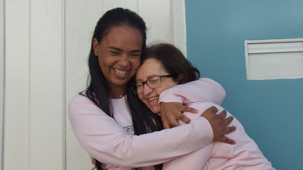 Na imagem, Claudiane e Martinha estão abraçadas e sorrindo. As duas vestem blusas rosa claro com o nome do projeto. Claudiane, mulher negra de cabelos longos, está à esquerda. Martinha, mulher branca de cabelos escuros e óculos, está à direita. Elas estão em frente a uma parede azul clara com uma porta branca ao lado.