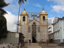 #paratodosverem: Fotografia da Igreja de Bom Jesus de Matozinhos, em Ouro Preto, em processo de restauração. A fachada da igreja, com duas torres amarelas está coberta por andaimes metálicos. À esquerda da imagem, há um canteiro de obras com chapas metálicas, materiais de construção empilhados e uma palmeira alta. À direita, há um prédio colonial de fachada branca com detalhes azuis e um operário de capacete caminhando próximo a pisos organizados no chão.