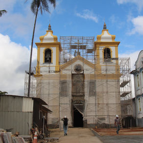 #paratodosverem: Fotografia da Igreja de Bom Jesus de Matozinhos, em Ouro Preto, em processo de restauração. A fachada da igreja, com duas torres amarelas está coberta por andaimes metálicos. À esquerda da imagem, há um canteiro de obras com chapas metálicas, materiais de construção empilhados e uma palmeira alta. À direita, há um prédio colonial de fachada branca com detalhes azuis e um operário de capacete caminhando próximo a pisos organizados no chão.