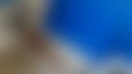 #ParaTodosVerem: Maca metálica antiga e enferrujada, instalada em um banheiro acessível com paredes de azulejos brancos. Sobre a maca, há dois colchonetes azuis dobrados e, ao lado, uma almofada com estampas coloridas. A superfície da maca apresenta sinais extensos de desgaste.