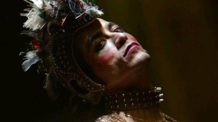 Cena do filme Homem com H com o ator Jesuíta Barbosa caracterizado como Ney Matogrosso. Ele aparece em um palco, usando um figurino chamativo com brilhos e penas na cabeça. A maquiagem é marcante, com batom vermelho e olhos destacados. Ele segura um microfone e olha diretamente para o público, em uma pose teatral e expressiva.