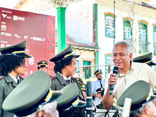 #PraTodosVerem: Na imagem, Efraim, Presidente da AMARBANDAS segura um microfone e conversa com dois jovens músicos da banda São Sebastião, ambos sorrindo. Os três estão cercados por músicos com fardas verde-oliva e boinas. Ao fundo, vê-se casarios históricos, árvores e o painel institucional do Festival Entrelaços.