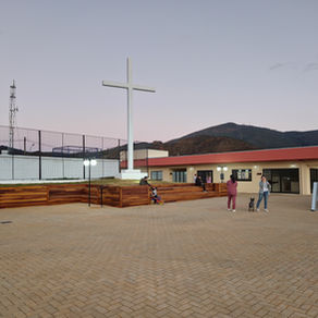 A imagem mostra o Parque Ecológico Estância do Cruzeiro, em Mariana (MG), ao entardecer. Em primeiro plano há uma ampla praça de piso claro. À esquerda ergue-se uma grande cruz branca sobre um pedestal, e à direita um prédio baixo de fachada clara e cobertura vermelha. Algumas pessoas caminham, conversam e uma delas passeia com um cachorro.