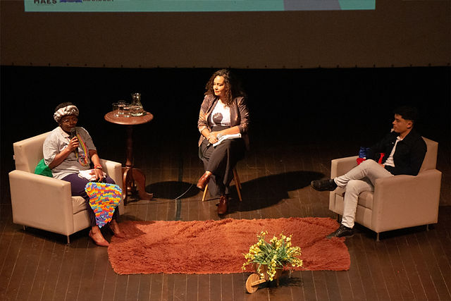 Três pessoas, cada uma em sua poltrona, em um palco, conversando no evento de fechamento da Semana da Diversidade.