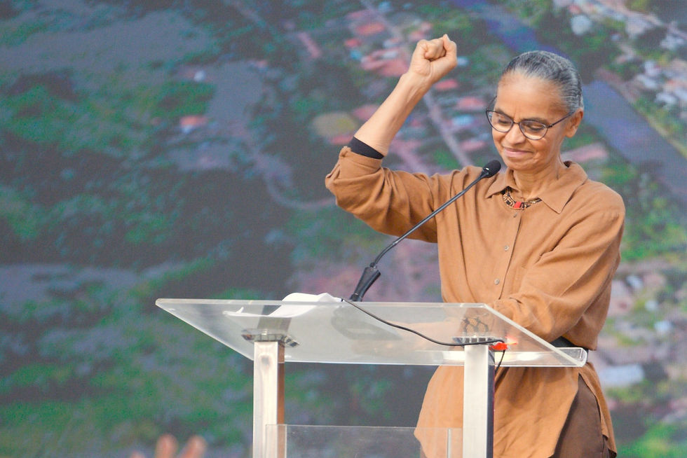 A imagem exibe a ministra Marina Silva, mulher negra de meia-idade, com cabelos presos e vestindo uma camisa marrom de mangas compridas, em um púlpito de acrílico. Ela está com o braço direito levantado e o punho cerrado, discursando. Ao fundo, um grande telão mostra uma paisagem aérea de um vale com um rio serpenteando e uma área urbana.