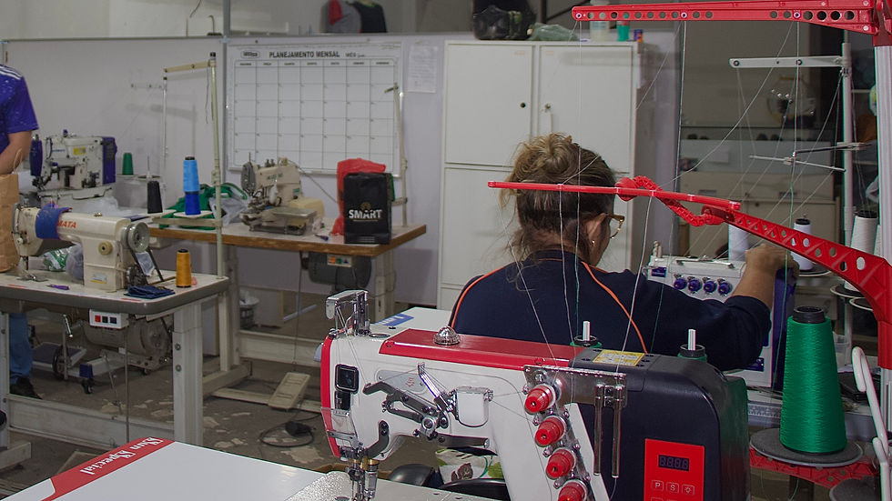 Na imagem, vemos o interior da Oficina de Corte & Costura. Em primeiro plano, uma mulher de costas, com cabelo preso e óculos, opera uma máquina de costura industrial. Há várias máquinas e linhas coloridas ao redor, e outras pessoas trabalhando ao fundo. Na parede, um quadro de planejamento mensal e armários brancos completam o ambiente.