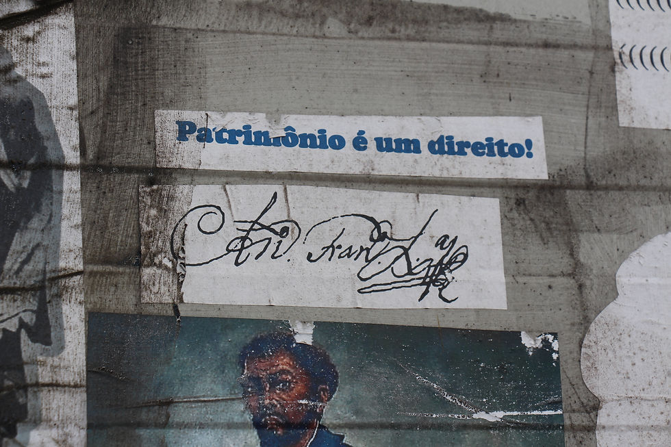 #paratodosverem Imagem de um painel desgastado, com a frase impressa “Patrimônio é um direito!” em letras azuis sobre fundo branco. Abaixo, há uma assinatura manuscrita antiga, em tinta preta, com traços cursivos e estilizados. O painel apresenta manchas e sinais de envelhecimento.