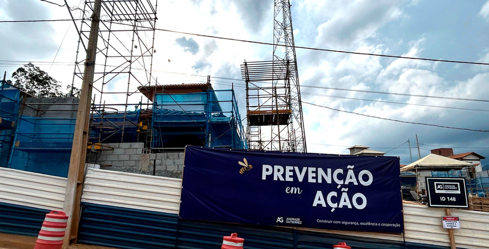#ParaTodosVerem: A imagem mostra uma obra em andamento, com elementos de construção como telas azuis e materiais de produção nas casas inacabadas. À frente, uma placa azul da construtora Andrade Gutierrez com letras brancas escritas: “Prevenção em Ação”. Abaixo, na mesma placa, está escrita a frase: “Construir com segurança, excelência e cooperação”.