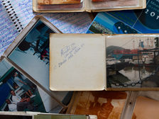 A imagem mostra uma composição de álbuns de fotografia antigos, fotos soltas e cadernos com anotações manuscritas, de forma sobrepostas. No centro, um pequeno álbum aberto revela uma página amarela, com uma assinatura, data e local. 