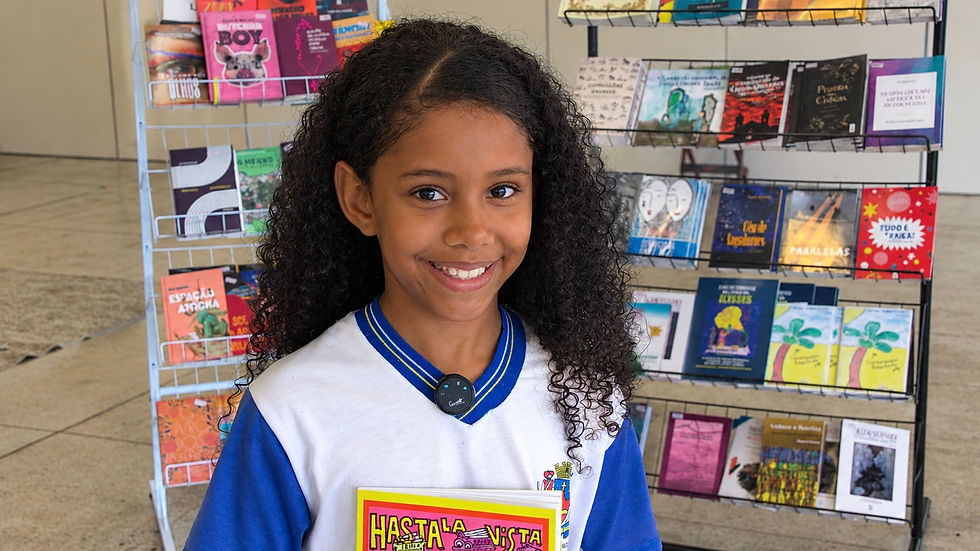 #ParaTodosVerem: Criança com cabelos cacheados, sorrindo. Ela veste um uniforme escolar branco com gola e manga azuis e segura um livro infantil rosa e amarelo. Ao fundo, há uma banca de livros infantis para venda.