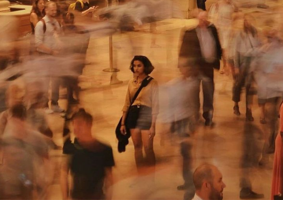 A person in a beige shirt stands still in a busy indoor setting. Blurred motion of people surrounds them, creating a sense of chaos.
