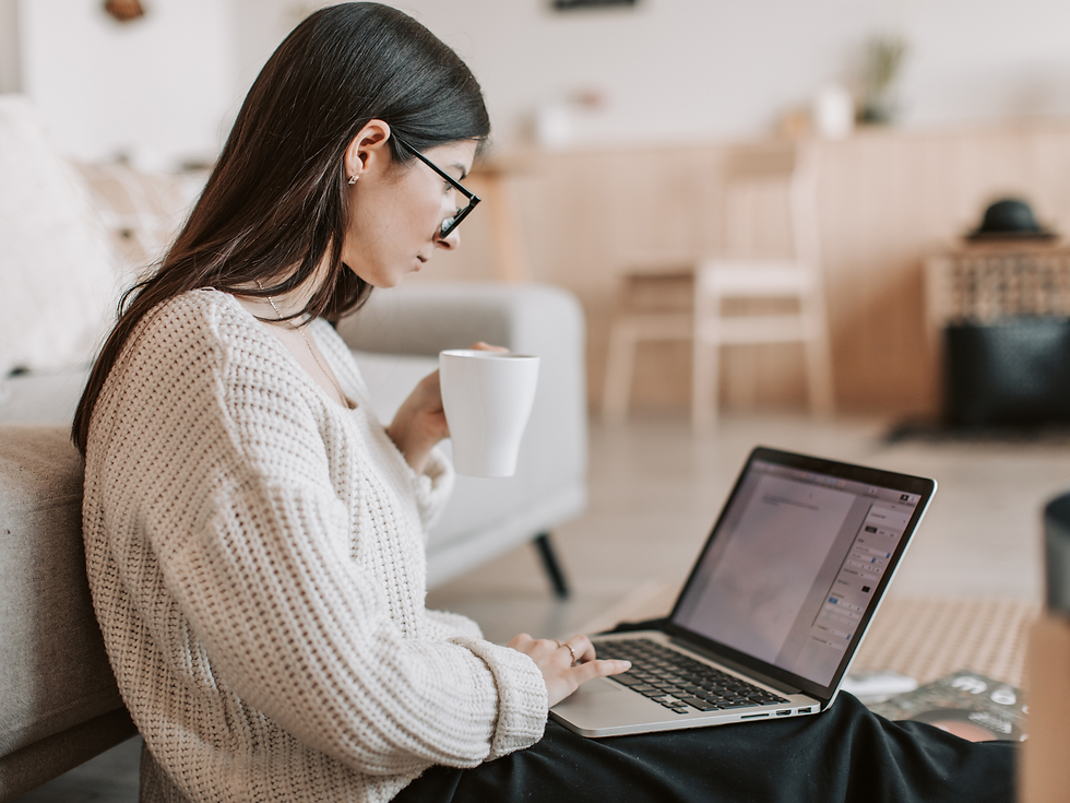 Woman in a cozy sweater sits on the floor, holding a mug, typing on a laptop in a softly lit room. Neutral tones create a calm mood.