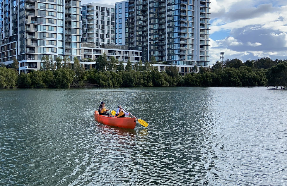 A mother and daughter paddle a canoe on the cooks river. The water has ripples and reflects the sky. There are aprtment buildings rising in the background.