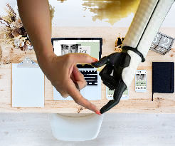 Human and robotic hands form a heart over a desk with a tablet, phone, clipboard, and flowers. Neutral colors dominate the scene.
