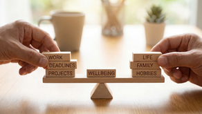 Two hands carefully balance wooden blocks on a seesaw, with “WORK,” “DEADLINES,” and “PROJECTS” stacked on one side, “LIFE,” “FAMILY,” and “HOBBIES” on the other, and “WELLBEING” centered in the middle on a table.