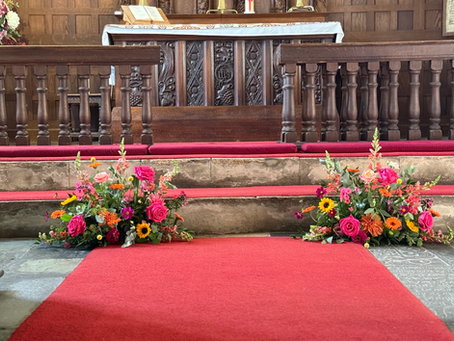 late summer wedding flowers arranged in front of the church alter, featuring sunflowers, cerise roses, Dahlias, and warm orange blooms