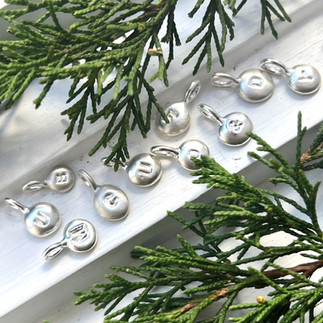 silver initial letter charms on a wooden white background with pine leaves