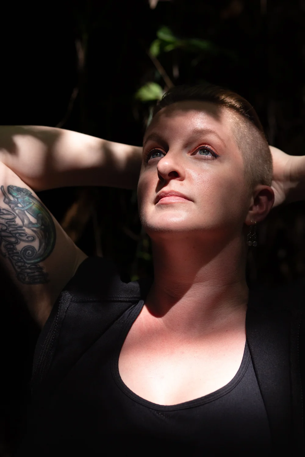 A headshot image of Sarah Gailey with short hair gazes upwards, arms behind head. Tattoo on arm visible. Sunlight filters through leaves, creating a serene mood.