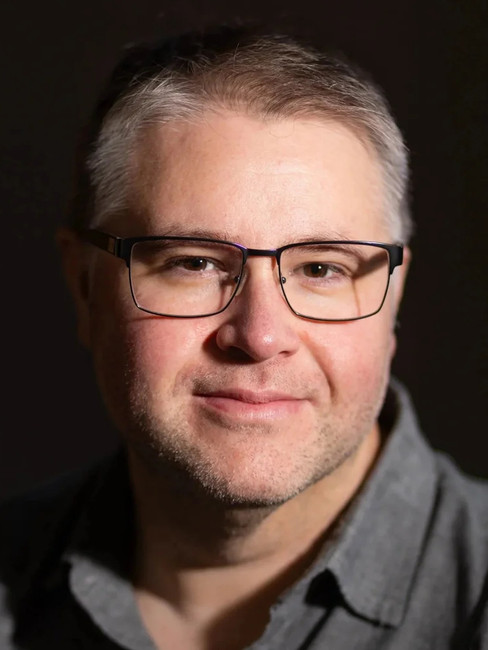 Close-up portrait of a middle-aged man with glasses and a slight smile against a dark background.