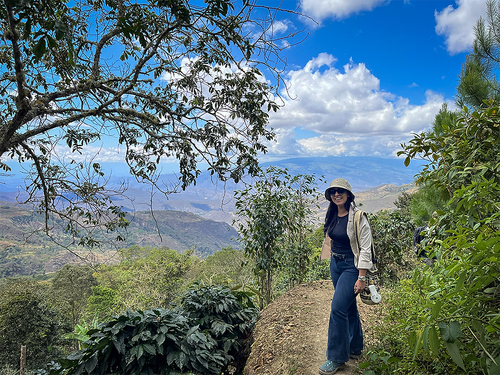 Smiling woman in hat stands on a lush mountain trail with valley views under a bright blue sky and clouds.