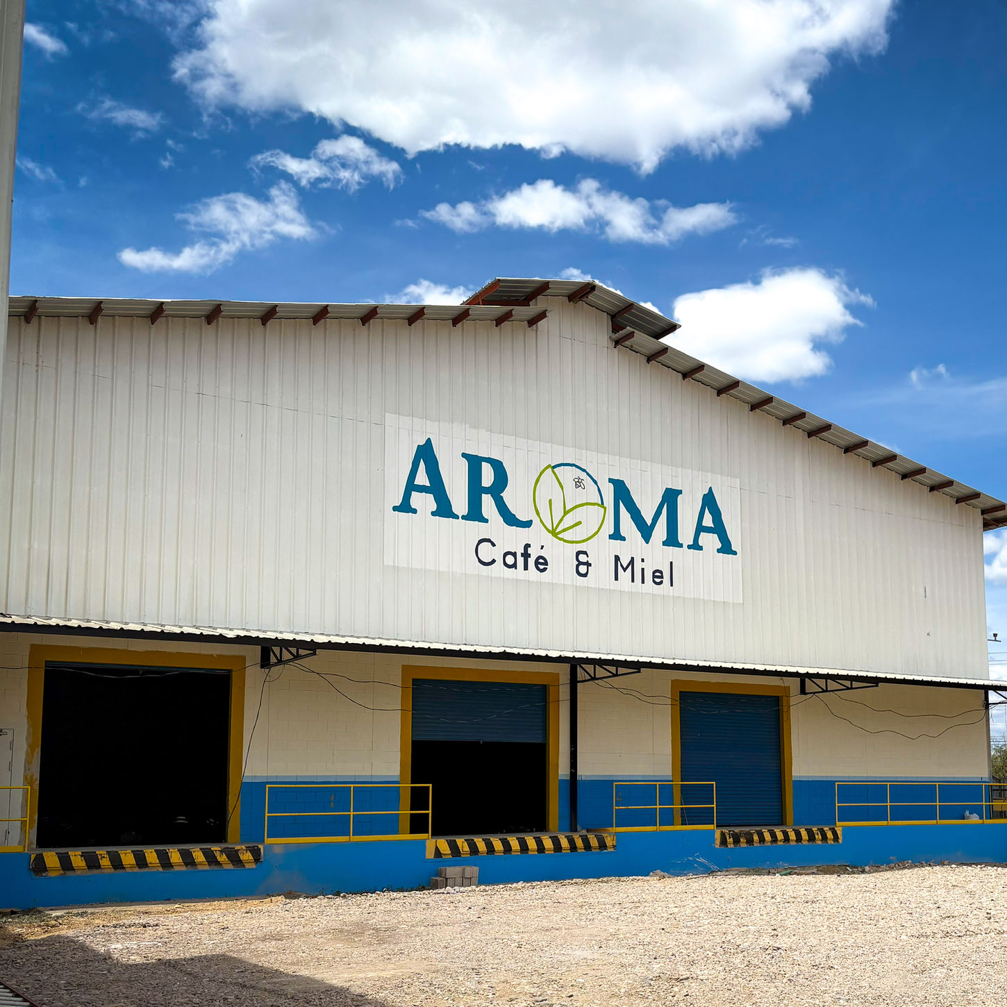 Exterior view of the ACM Coffee Processing Mill, showing the Aroma Café & Miel building under a bright blue sky with scattered clouds.