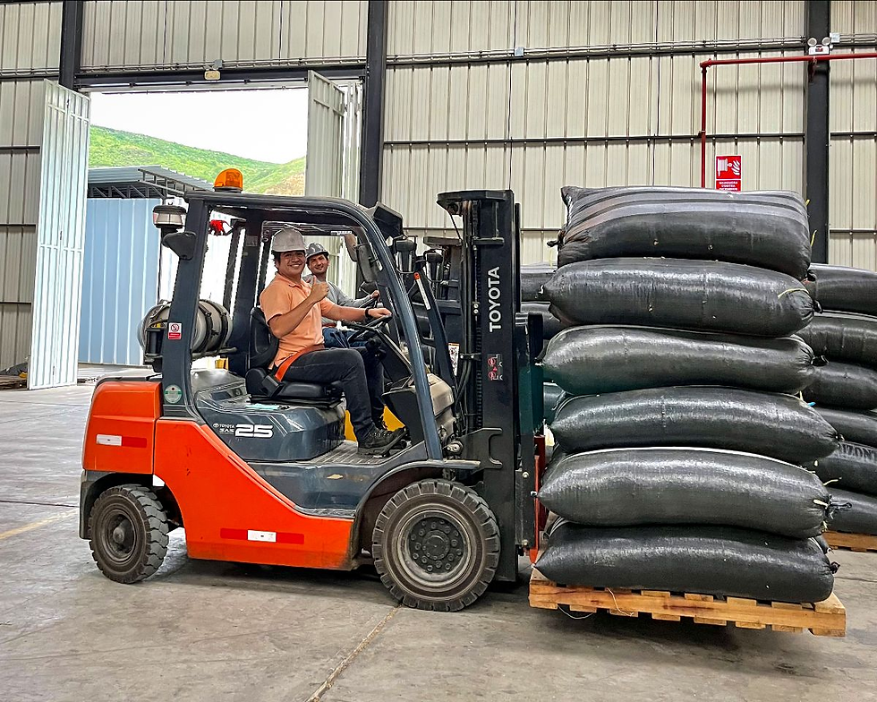 Two people on an orange forklift in a warehouse, smiling. Black bags stacked on a pallet. Open doors reveal green hills outside.