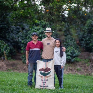 Three people smiling beside a coffee sack labeled "Excelso Café de Colombia." They're in a lush, green coffee farm setting.