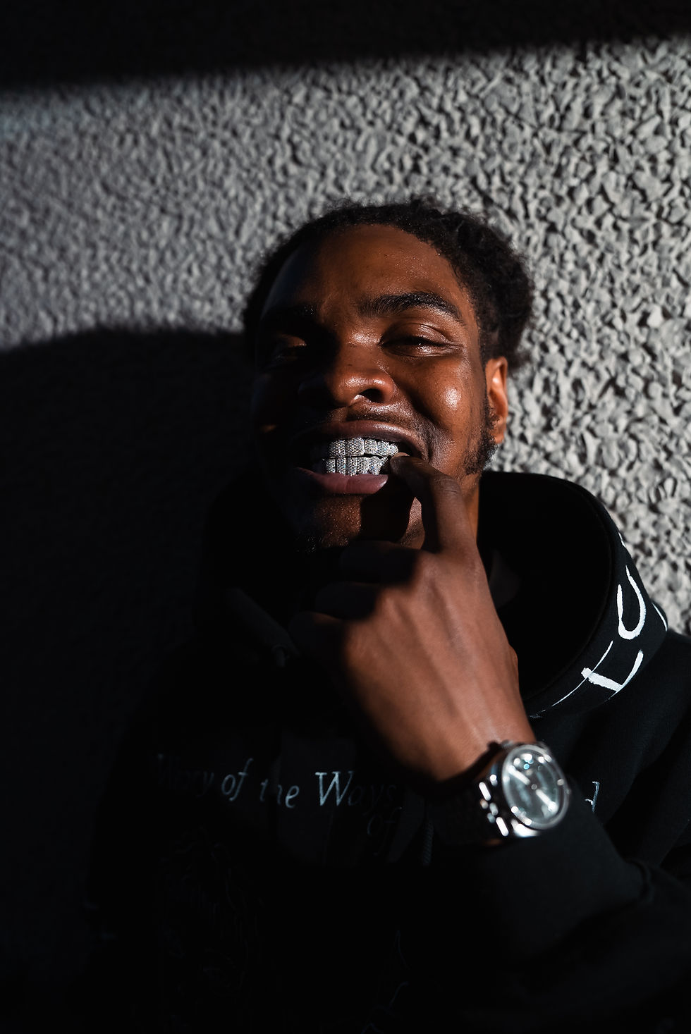 Man with a big smile shows his grillz, wearing a black hoodie, silver watch, against textured wall in shadow. Playful and confident mood.