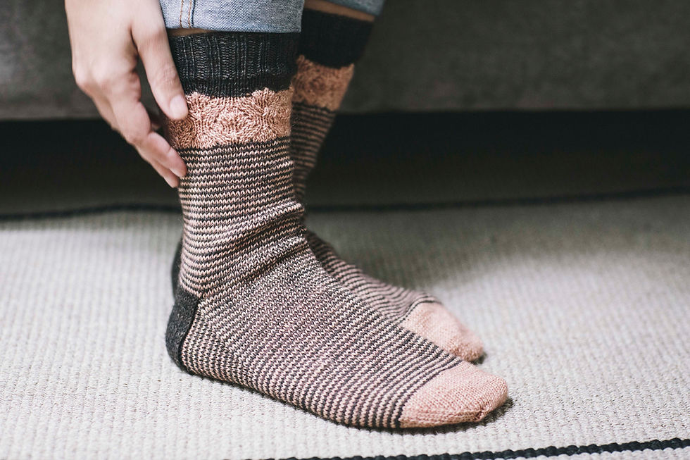 Thumbnail: 2 feet wearing hand-knit socks in pink and black. Hands reaching the ankles. The background is a gray velvet sofa.