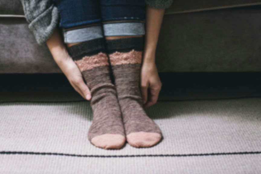 2 feet wearing hand-knit socks in pink and black. Hands reaching the ankles. The background is a gray velvet sofa.
