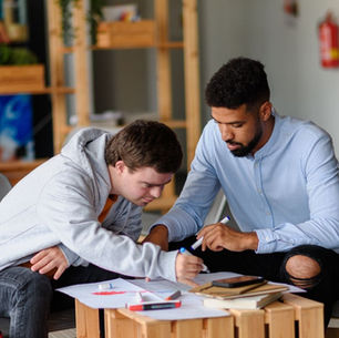 A young man in a gray hoodie writes on paper with a marker while a man in a blue striped shirt sits beside him and points to the page, at a low wooden table.