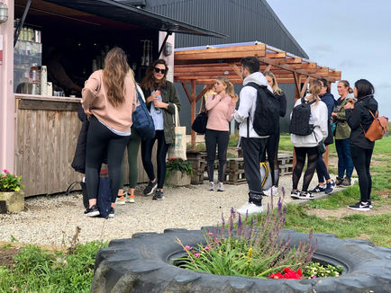 People in casual athletic wear gathered at a rustic outdoor refreshment stand with wooden counter and flower-filled tire planter.