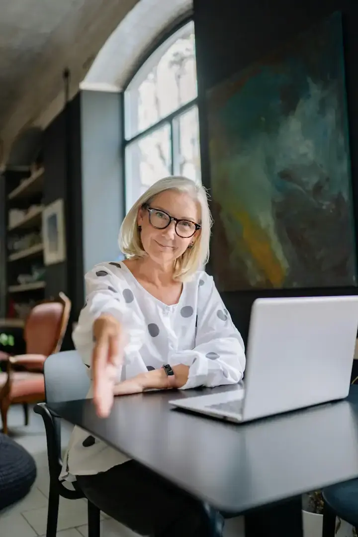 Mujer mayor sonriente con gafas ofrece mano, sentada con portátil.