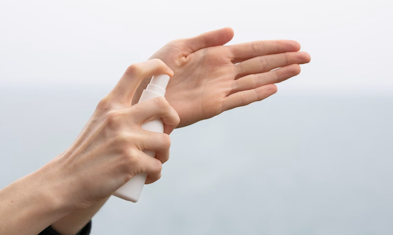 Hands using a spray bottle to spritz liquid onto the palm, against a blurred blue and white background. Calm, simple, with focus on hands.