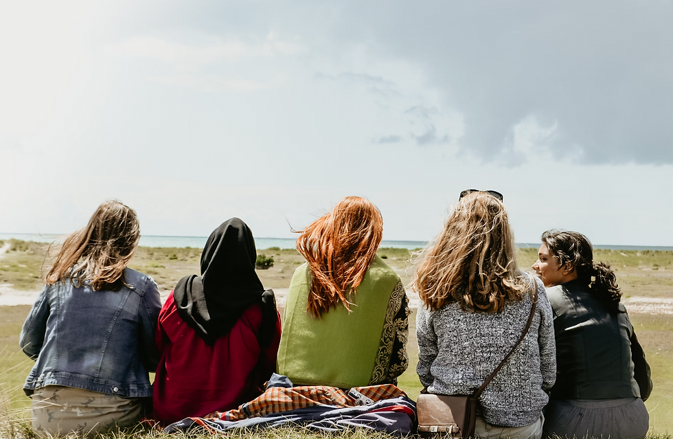 Five people sit on grass, backs to the camera, facing the ocean. Wind blows their hair. Sky is partly cloudy. Day feels relaxed.