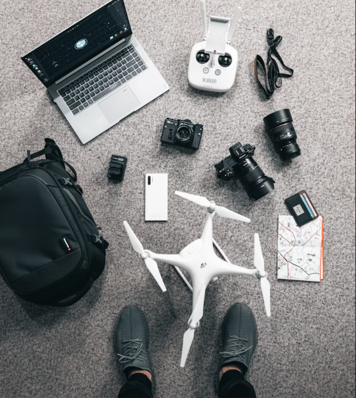 Drone, camera, laptop, and travel gear neatly arranged on carpet. Person's feet visible. Map and passport suggest travel. Neutral colors.