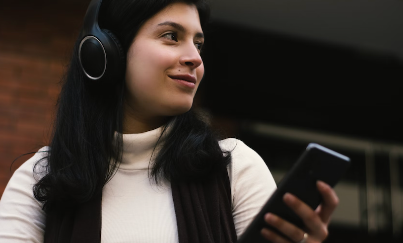 Woman with dark hair wearing headphones, smiling and holding a smartphone. She is outdoors with a blurred brick wall in the background.