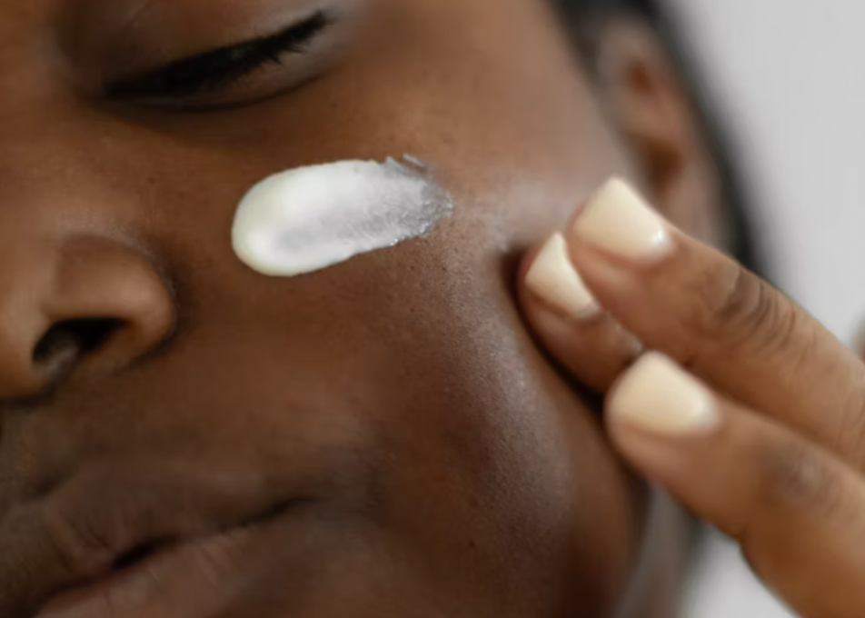 Close-up of a person applying white cream on their cheek with fingers. Skin tone is warm. Neutral background, focus on skincare routine.