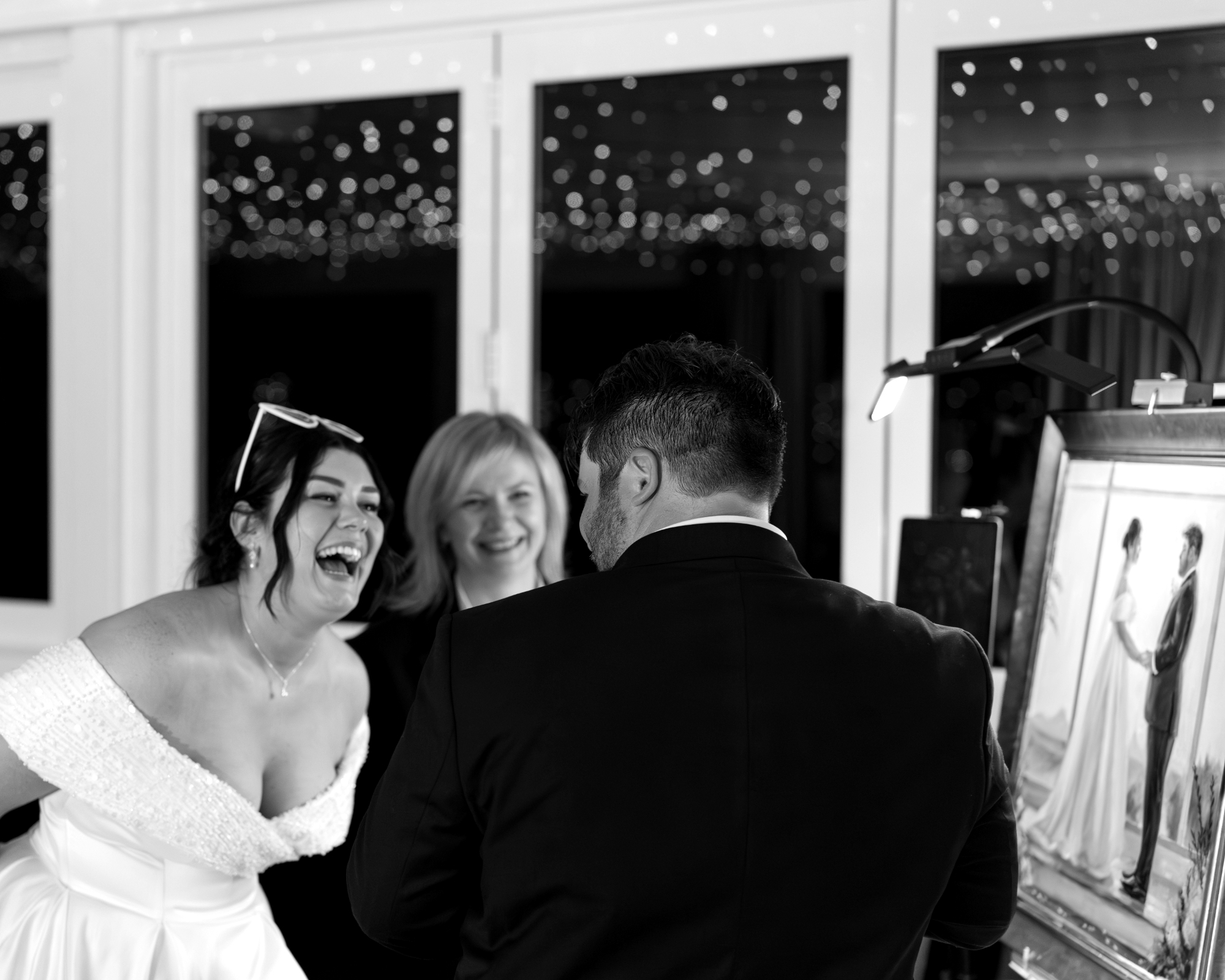 A joyful bride, Caitlyn, and groom, Jake, stand beside artist Elena Valerie, their faces filled with happiness as they look upon their completed live wedding painting, displayed on an easel during their reception at Tiffany's Maleny.