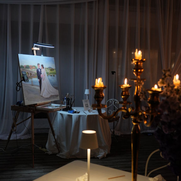 Live wedding painting setup featuring an oil painting of a bride and groom on an easel, illuminated by an artist's lamp, with a candelabra and art supplies on a nearby table at an elegant event
