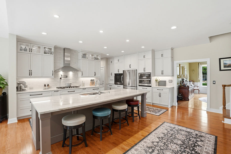 Modern white kitchen featuring a spacious waterfall island, custom shaker cabinetry, quartz countertops, stainless steel appliances, and open-concept design with hardwood floors.