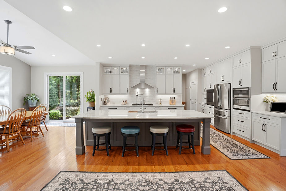 Front view of a luxury kitchen renovation showcasing a large island with seating, white shaker cabinets, stainless steel range hood, quartz countertops, and an open floor plan.
