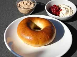Bagel on a white plate with bowls of whipped butter and berries on a gray surface. Bright, fresh setting with a focus on textures.