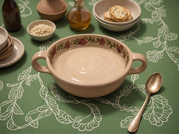 Tahini sauce in a floral bowl on a green tablecloth. Surrounding are bowls, olive oil, and a spoon. Cozy, inviting atmosphere.