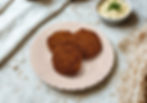 Three breaded patties on a pink plate, accompanied by a bowl of creamy dip garnished with herbs. Soft white fabric and netting nearby.