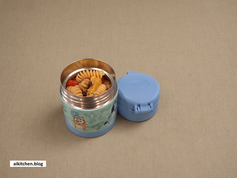 Blue lidded thermos with pasta salad, featuring veggies, on a beige surface. Cartoon animals on the thermos. Text: aikitchen.blog.