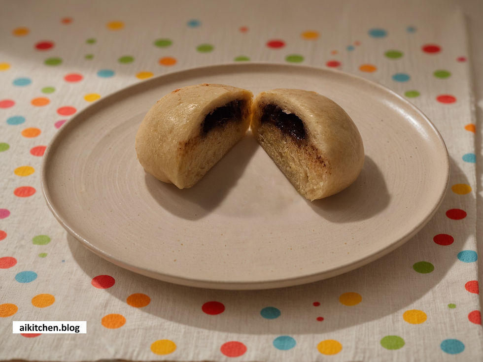 A sliced steamed bun on a beige plate reveals dark filling, set on a colorful polka dot tablecloth. "aikitchen.blog" is visible.